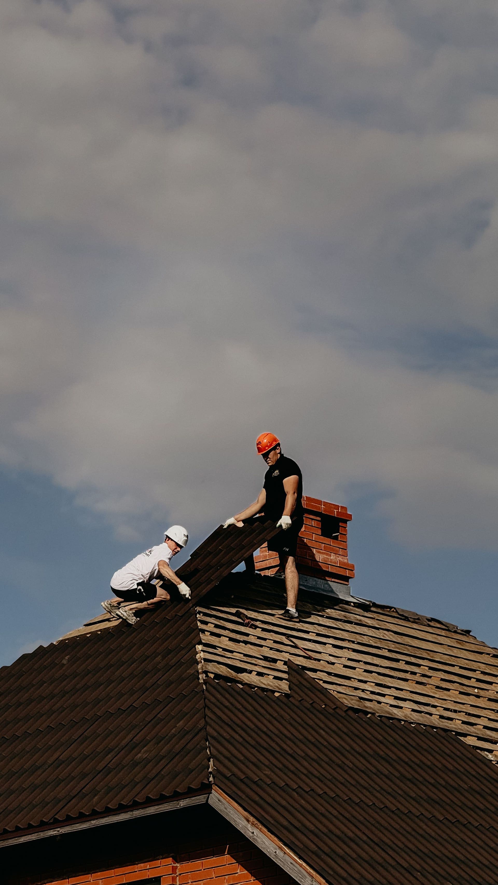 Two workers on a residential roof beside a red brick chimney